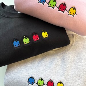 May include: Three sweatshirts in different colors, pink, black, and gray, with embroidered pixelated characters in blue, green, red, and yellow.