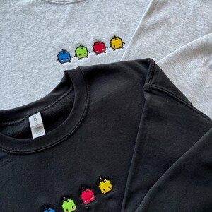 May include: Two sweatshirts with embroidered pixelated characters. The top sweatshirt is gray with five characters in blue, green, red, yellow, and yellow. The bottom sweatshirt is black with five characters in blue, green, red, yellow, and yellow.