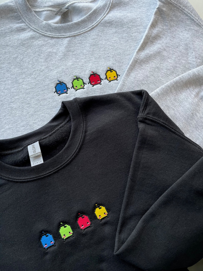 May include: Two sweatshirts with embroidered pixelated characters. The top sweatshirt is gray with five characters in blue, green, red, yellow, and yellow. The bottom sweatshirt is black with five characters in blue, green, red, yellow, and yellow.