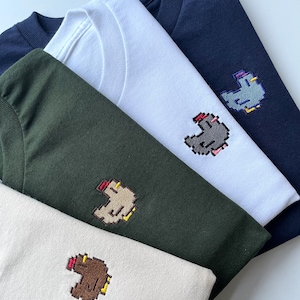 May include: Four t-shirts in different colors with a pixelated chicken design embroidered on the front. The shirts are stacked on top of each other, with the navy blue shirt on top and the beige shirt on the bottom.