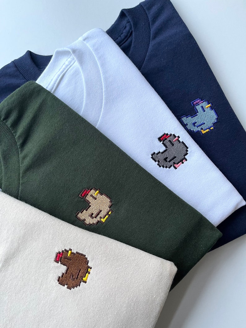May include: Four t-shirts in different colors with a pixelated chicken design embroidered on the front. The shirts are stacked on top of each other, with the navy blue shirt on top and the beige shirt on the bottom.