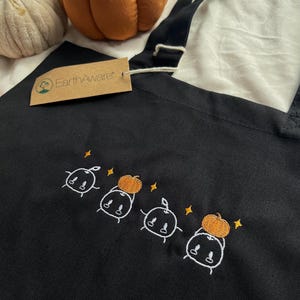 Op de afbeelding: Een zwarte draagtas met een geborduurd ontwerp van vier witte, geestachtige figuren, waarvan er twee oranje pompoenen vasthouden, geaccentueerd door kleine gele sterren. Een bruin kartonnen label met de tekst EarthAware zit aan de riem.