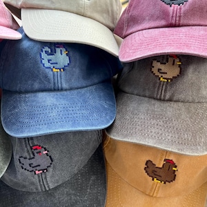 May include: A collection of six baseball caps in various colors including beige, maroon, blue, grey, and tan. Each cap features a small, embroidered pixel art chicken design on the front panel. The hats have a washed, vintage-style fabric texture.