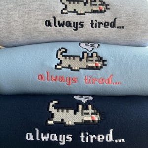 May include: Three sweatshirts stacked on top of each other. The sweatshirts are gray, light blue, and navy blue. Each sweatshirt has a pixelated cat with a thought bubble above its head that says "zz" and the text "always tired..." embroidered on the front.
