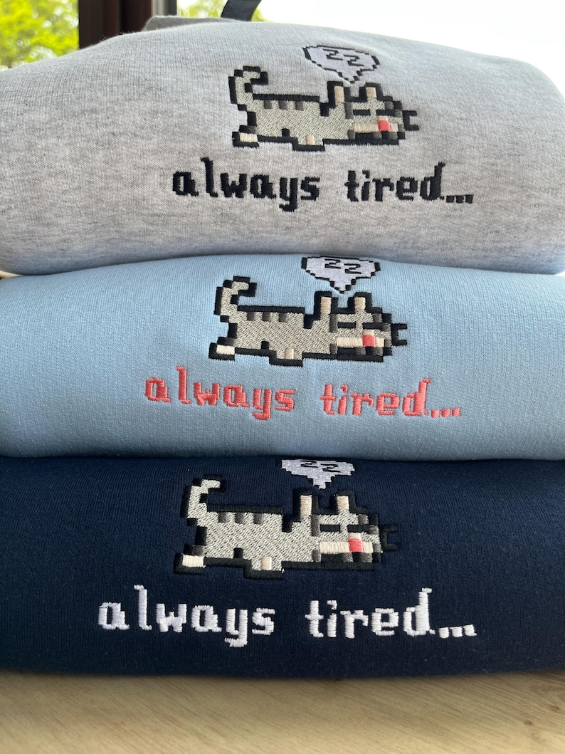 May include: Three sweatshirts stacked on top of each other. The sweatshirts are gray, light blue, and navy blue. Each sweatshirt has a pixelated cat with a thought bubble above its head that says "zz" and the text "always tired..." embroidered on the front.