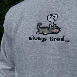 May include: A gray long-sleeved sweatshirt with a pixelated cat design and the text "always tired..." in black embroidery. The cat is holding a speech bubble with the letters "zz" inside.