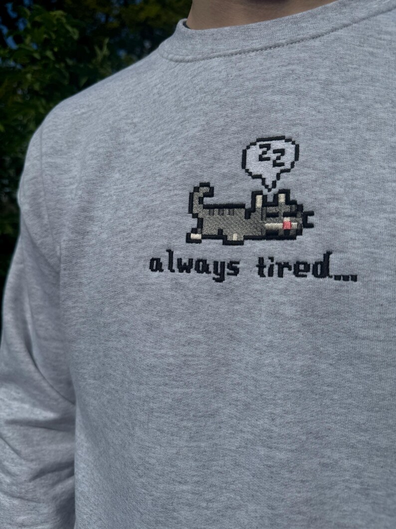 May include: A gray long-sleeved sweatshirt with a pixelated cat design and the text "always tired..." in black embroidery. The cat is holding a speech bubble with the letters "zz" inside.
