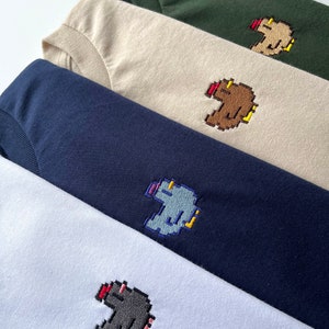 Puede incluir: Cuatro camisetas de diferentes colores, cada una con una imagen bordada pixelada de una criatura con la nariz roja. Los colores de las camisetas son blanco, azul marino, beige y verde bosque.