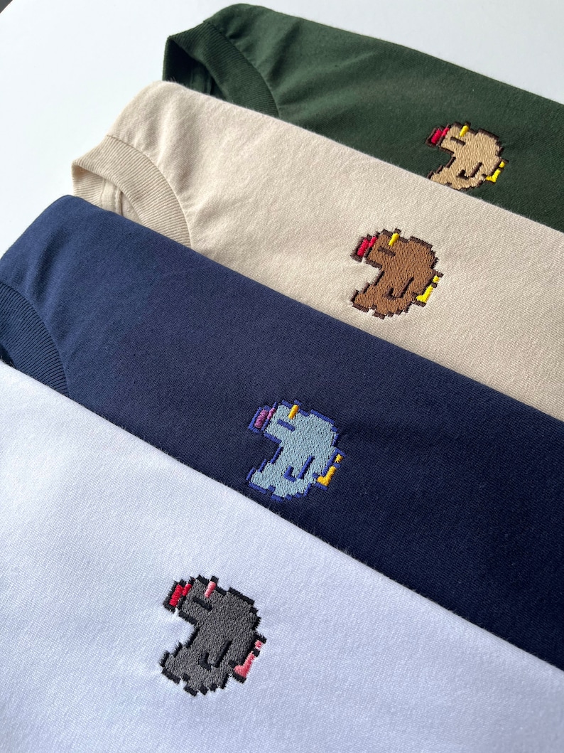 May include: Four t-shirts in different colors, each with a pixelated embroidered image of a creature with a red nose. The colors of the shirts are white, navy blue, beige, and forest green.