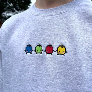 May include: A gray sweatshirt with a row of four embroidered pixelated characters. The characters are blue, green, red, and yellow and have smiling faces.