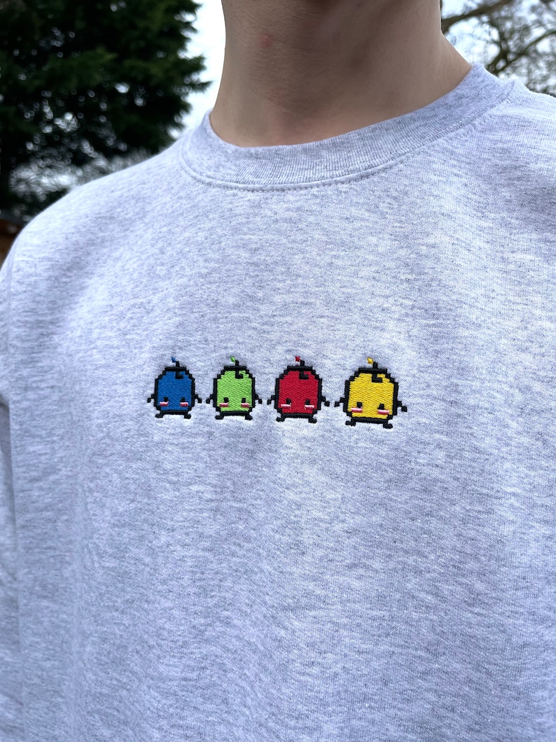May include: A gray sweatshirt with a row of four embroidered pixelated characters. The characters are blue, green, red, and yellow and have smiling faces.
