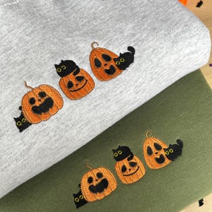 May include: Two sweatshirts with embroidered black cats and orange pumpkins. The top sweatshirt is grey and the bottom sweatshirt is green.