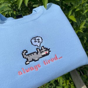 May include: Light blue sweatshirt with a pixelated cat design and the text "zzz" in a speech bubble above the cat. The text "always tired..." is embroidered below the cat.