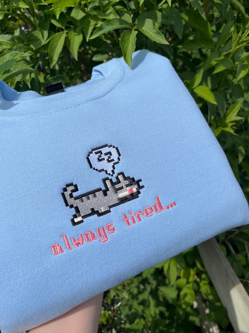 May include: Light blue sweatshirt with a pixelated cat design and the text "zzz" in a speech bubble above the cat. The text "always tired..." is embroidered below the cat.