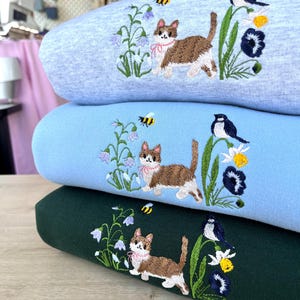 Puede incluir: Tres sudaderas apiladas una encima de la otra. La sudadera de arriba es azul claro con un gato, flores y una abeja bordadas. La sudadera del medio es azul claro con un gato, flores y una abeja bordadas. La sudadera de abajo es verde oscuro con un gato, flores y una abeja bordadas.