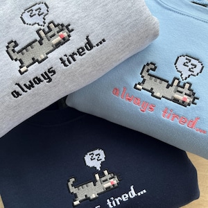 May include: Three sweatshirts with a pixelated cat design and the text "always tired..."  The sweatshirts are in grey, blue, and navy blue.