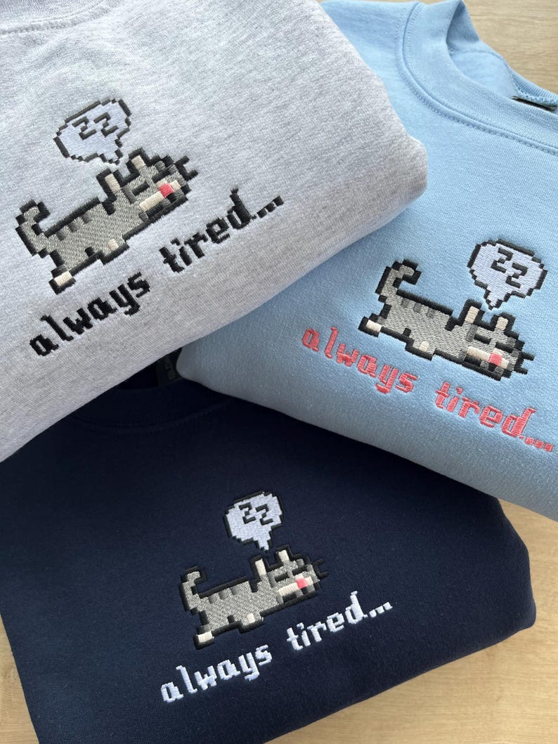 May include: Three sweatshirts with a pixelated cat design and the text "always tired..."  The sweatshirts are in grey, blue, and navy blue.