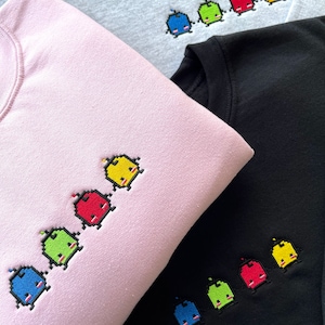 Könnte beinhalten: Drei Sweatshirts in verschiedenen Farben mit einer Reihe von gestickten Pixelfiguren. Die Figuren sind blau, grün, rot und gelb. Die Sweatshirts sind grau, rosa und schwarz.