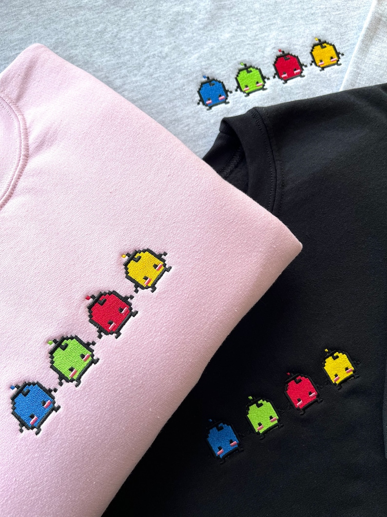 May include: Three sweatshirts in different colors with a line of embroidered pixelated characters. The characters are blue, green, red, and yellow. The sweatshirts are gray, pink, and black.