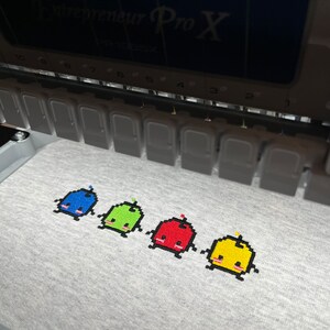 May include: Four embroidered pixelated characters on a gray fabric. The characters are blue, green, red, and yellow. Each character has a small, round head, a body, and two small legs. The characters are arranged in a row from left to right.