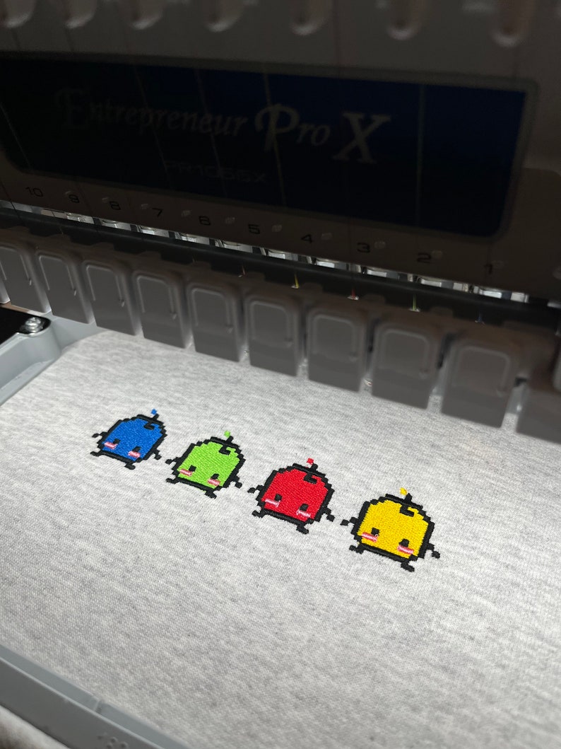 May include: Four embroidered pixelated characters on a gray fabric. The characters are blue, green, red, and yellow. Each character has a small, round head, a body, and two small legs. The characters are arranged in a row from left to right.