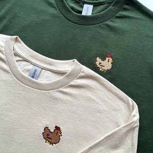 May include: Two t-shirts with embroidered pixelated chickens. One t-shirt is a light beige color and the other is a dark green color. Both shirts have the Gildan brand tag.