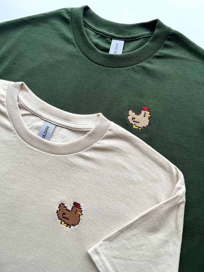May include: Two t-shirts with embroidered pixelated chickens. One t-shirt is a light beige color and the other is a dark green color. Both shirts have the Gildan brand tag.