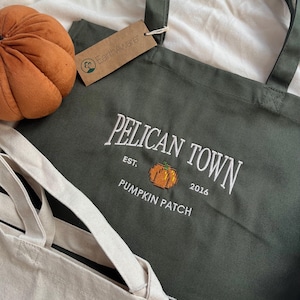 May include: A dark olive green canvas tote bag featuring white embroidered text that reads PELICAN TOWN EST. 2016 PUMPKIN PATCH with a small orange pixelated pumpkin graphic. An EarthAware brand tag is attached to the handle.