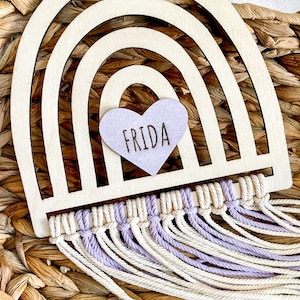 Może przedstawiać: Drewniany wiszący tęcza z fioletowym sercem, na którym widnieje napis "FRIDA". Tęcza ma frędzle z makramą z białej i fioletowej przędzy.