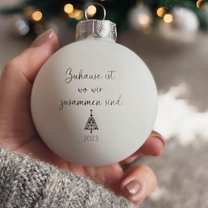 May include: A matte white spherical Christmas bauble featuring the black printed text "Zuhause ist, wo wir zusammen sind," a small stylized Christmas tree graphic composed of stars, and the year "2023" printed in black at the bottom.