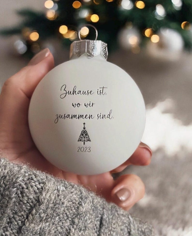 May include: A matte white spherical Christmas bauble featuring the black printed text "Zuhause ist, wo wir zusammen sind," a small stylized Christmas tree graphic composed of stars, and the year "2023" printed in black at the bottom.