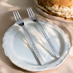 May include: Two silver forks with the inscription "Friedrich dit merci." on a white plate. The forks are on a light pink surface.