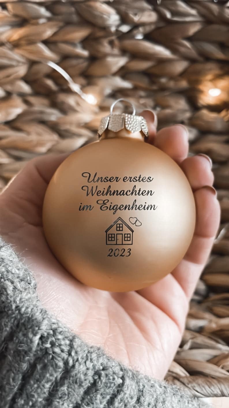 May include: A matte gold spherical Christmas bauble featuring the black printed text "Unser erstes Weihnachten im Eigenheim," a small house icon with two hearts, and the year "2023" centred on the front surface with a silver metal cap.
