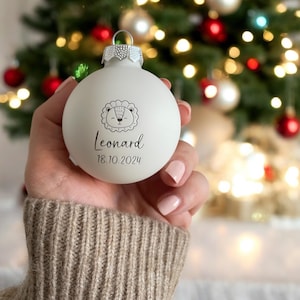 May include: A matte white spherical Christmas bauble featuring a black line drawing of a lion face, the name Leonard, and the date 18.10.2024 printed in black text. The bauble has a silver metal cap with a small loop for hanging.