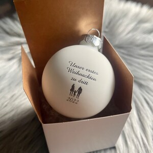 May include: A matte white spherical Christmas bauble featuring black printed text that reads "Unser erstes Weihnachten zu dritt" above a silhouette of two adults and a child, with the year "2022" printed below the figures, housed in a box.