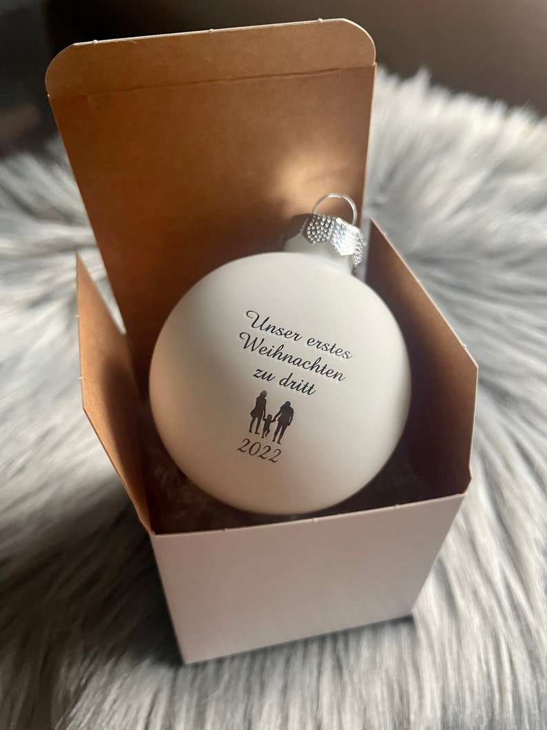 May include: A matte white spherical Christmas bauble featuring black printed text that reads "Unser erstes Weihnachten zu dritt" above a silhouette of two adults and a child, with the year "2022" printed below the figures, housed in a box.