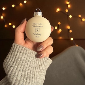 May include: A matte cream-coloured spherical Christmas bauble featuring the printed text Mein erstes Weihnachten, a small lion illustration, the name Ludwig, and the year 2025 in black ink, topped with a silver-toned metal cap and hanging loop.
