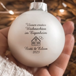 May include: A matte white spherical Christmas bauble featuring the black printed text "Unser erstes Weihnachten im Eigenheim", a small house icon with two hearts, and the names "Botti & Eileen" followed by the year "2023" in a serif font.
