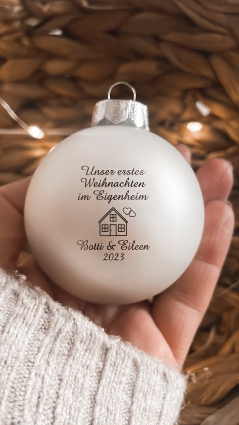 May include: A matte white spherical Christmas bauble featuring the black printed text "Unser erstes Weihnachten im Eigenheim", a small house icon with two hearts, and the names "Botti & Eileen" followed by the year "2023" in a serif font.