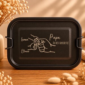 May include: A rectangular black lunch box with rounded corners and two side metal clips. The lid features a laser-engraved design of three fists bumping together, with the names Luana and Eliano, and the text Papa DU BIST DER BESTE!