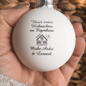 May include: A matte white spherical Christmas bauble featuring black printed text that reads "Unser erstes Weihnachten im Eigenheim", the year "2024", a small house icon with two hearts, and the names "Maike, Andr&eacute; & Leonard".