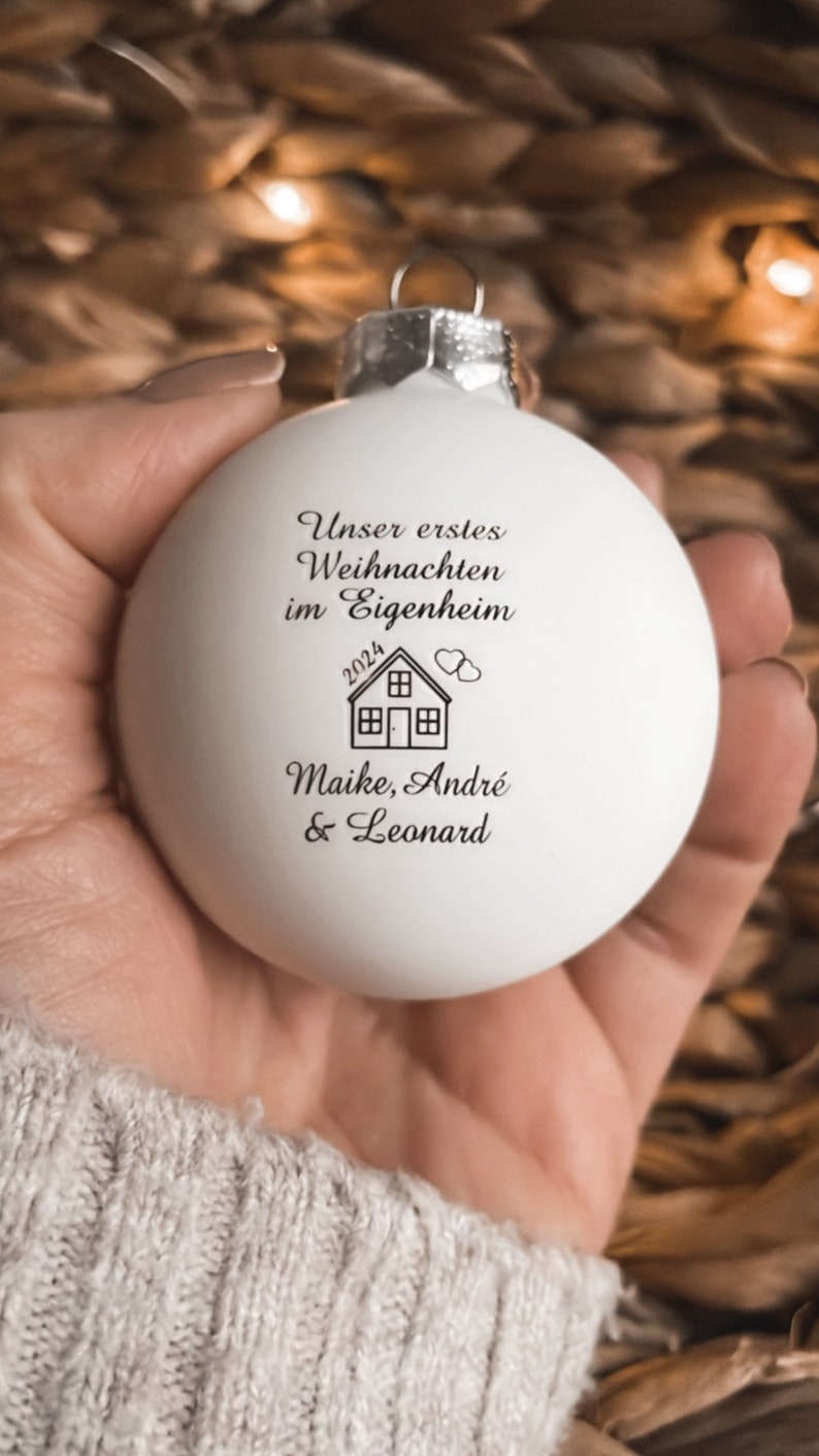 May include: A matte white spherical Christmas bauble featuring black printed text that reads "Unser erstes Weihnachten im Eigenheim", the year "2024", a small house icon with two hearts, and the names "Maike, Andr&eacute; & Leonard".