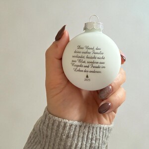 May include: A matte white spherical Christmas ornament featuring black printed German text that reads: Das Band, das deine wahre Familie verbindet, besteht nicht aus Blut, sondern aus Respekt und Freude im Leben des anderen. Includes a small tree icon and 2025.