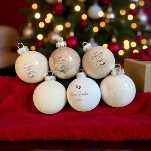 May include: A collection of six round, smooth Christmas baubles in shades of cream and white. Three baubles feature a black line drawing of hands forming a heart with the text Unser Weihnachten zu viert. One bauble displays baby footprints and the text Kleines Wunder 2025.