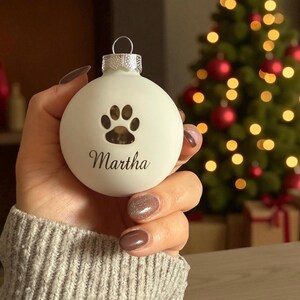 May include: A matte white spherical Christmas bauble featuring a cutout paw print design above the name Martha in black script lettering. The bauble has a silver metal cap with a small loop at the top for hanging on a tree branch.