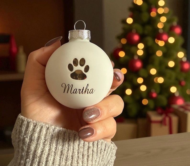 May include: A matte white spherical Christmas bauble featuring a cutout paw print design above the name Martha in black script lettering. The bauble has a silver metal cap with a small loop at the top for hanging on a tree branch.