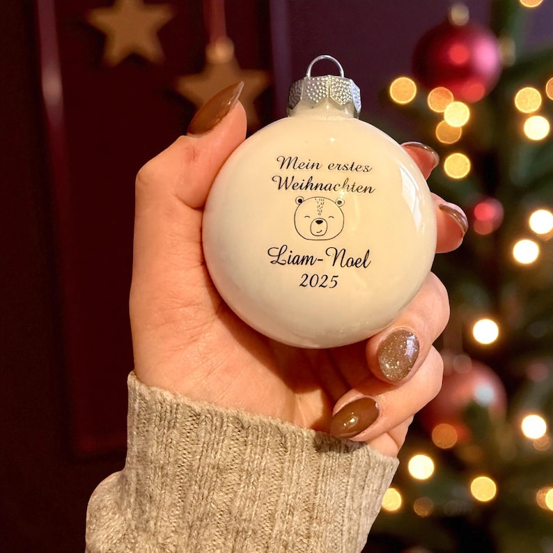 May include: A white spherical Christmas bauble featuring a silver metal cap. The bauble is printed with the text Mein erstes Weihnachten, a line drawing of a bear face, the name Liam-Noel, and the year 2025 in a dark script font.