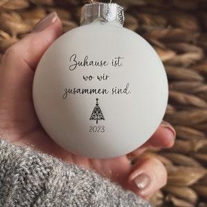 May include: A matte white spherical Christmas bauble with a silver metal cap. The front features black text that reads "Zuhause ist, wo wir zusammen sind." above a small black graphic of a Christmas tree and the year "2023" printed below.