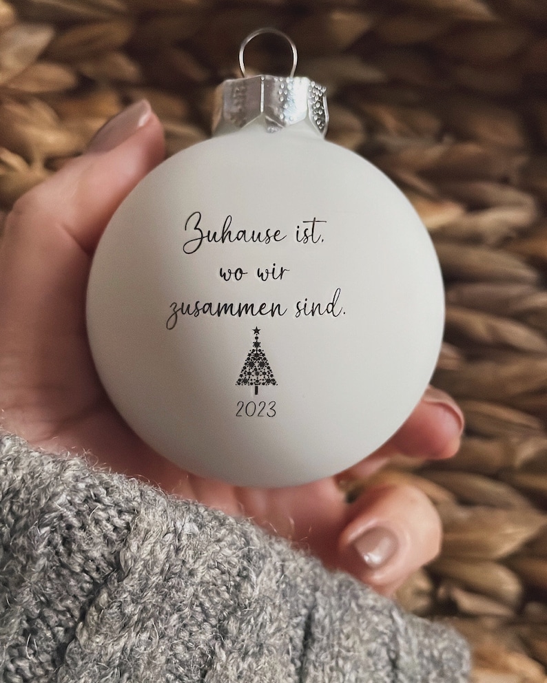May include: A matte white spherical Christmas bauble with a silver metal cap. The front features black text that reads "Zuhause ist, wo wir zusammen sind." above a small black graphic of a Christmas tree and the year "2023" printed below.