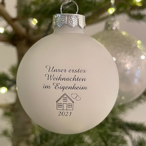 May include: A matte white spherical Christmas bauble with a silver cap. The front features black printed text that reads "Unser erstes Weihnachten im Eigenheim" above a line drawing of a house with two small hearts and the year 2021 below.